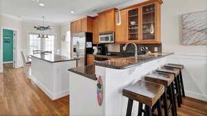 A modern kitchen with wooden cabinets, a central island with bar stools, stainless steel appliances, and pendant lighting. - Whalecome to the beach-elevator (Carolina Beach)
