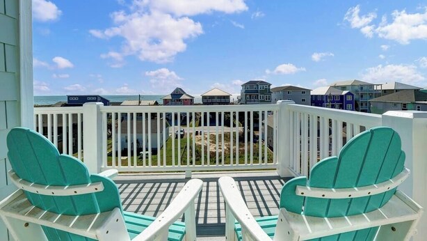 A balcony with two turquoise Adirondack chairs, a white railing, and a view of the ocean and beach houses. - Whalecome to the beach-elevator (Carolina Beach)