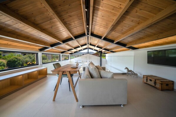 A spacious living area with a high ceiling, wooden beams, a sofa, a dining table, and a view of greenery outside.