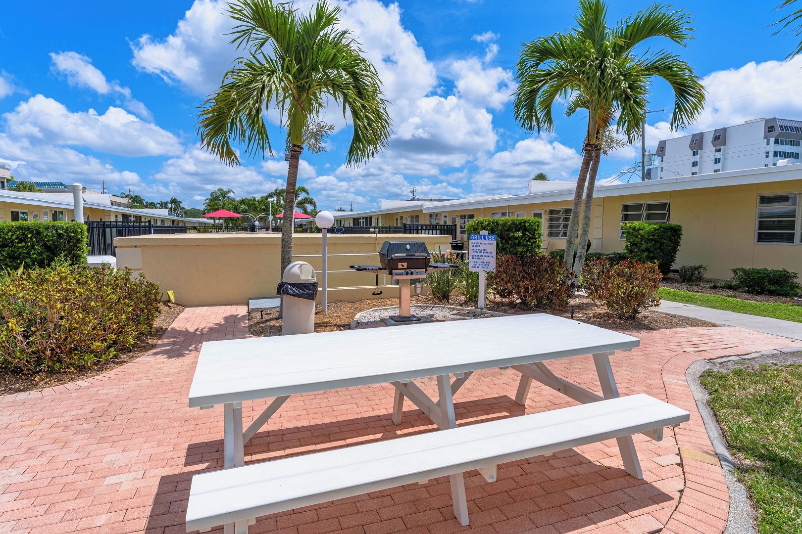 A picnic table with a grill in a courtyard with palm trees and a building in the background.
