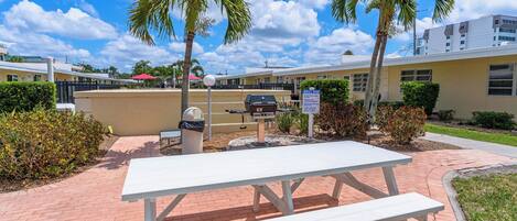 A picnic table with a grill in a courtyard with palm trees and a building in the background.