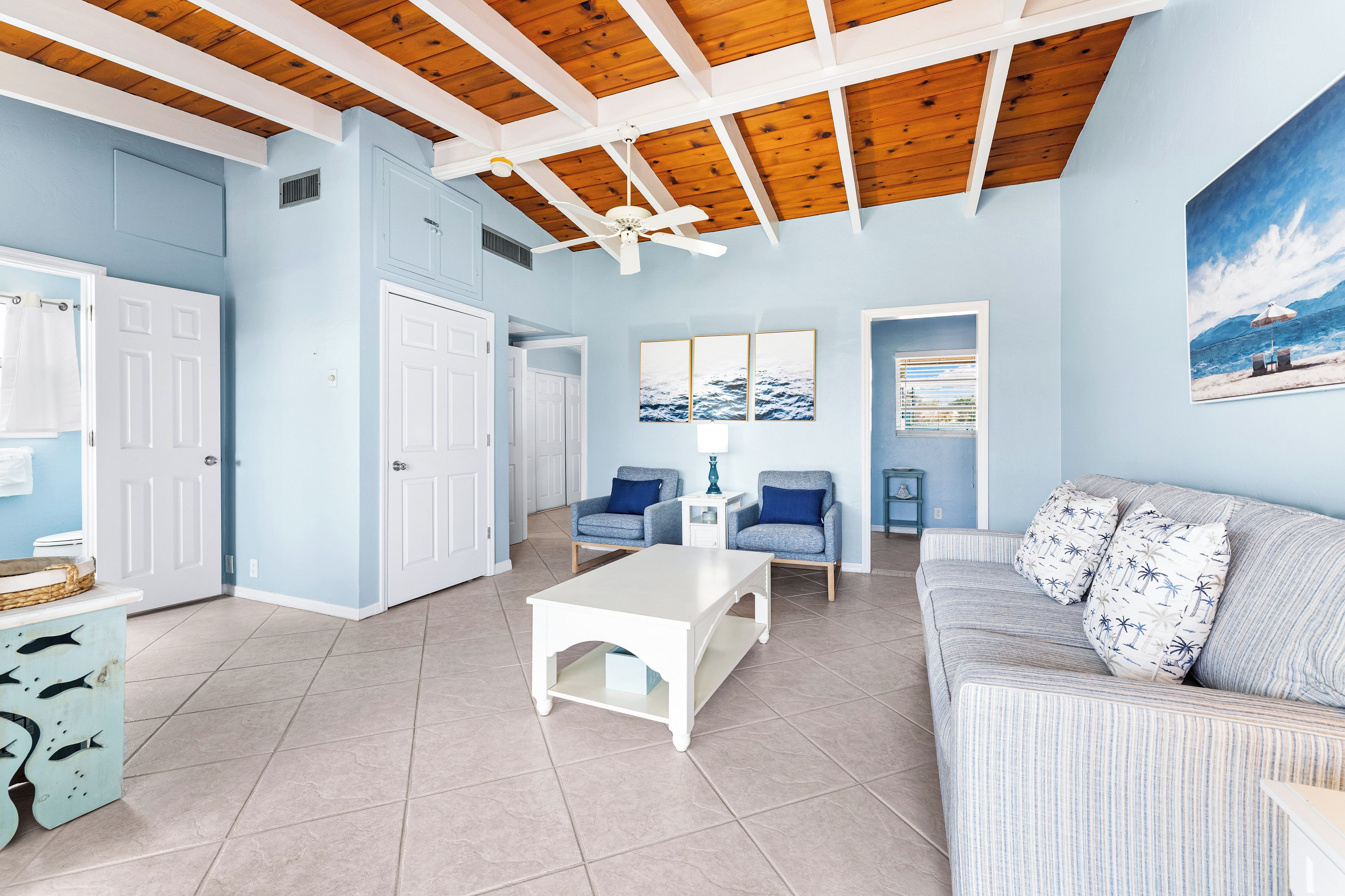 Un salon avec une table basse blanche, deux fauteuils bleus, un canapé à rayures et un ventilateur de plafond.