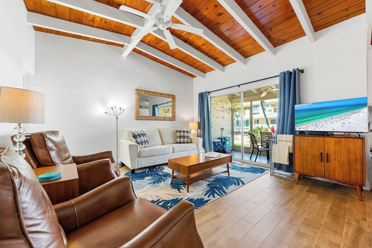 A living room with a sofa, coffee table, and TV, wooden ceiling beams, and a view of the beach through a glass door.