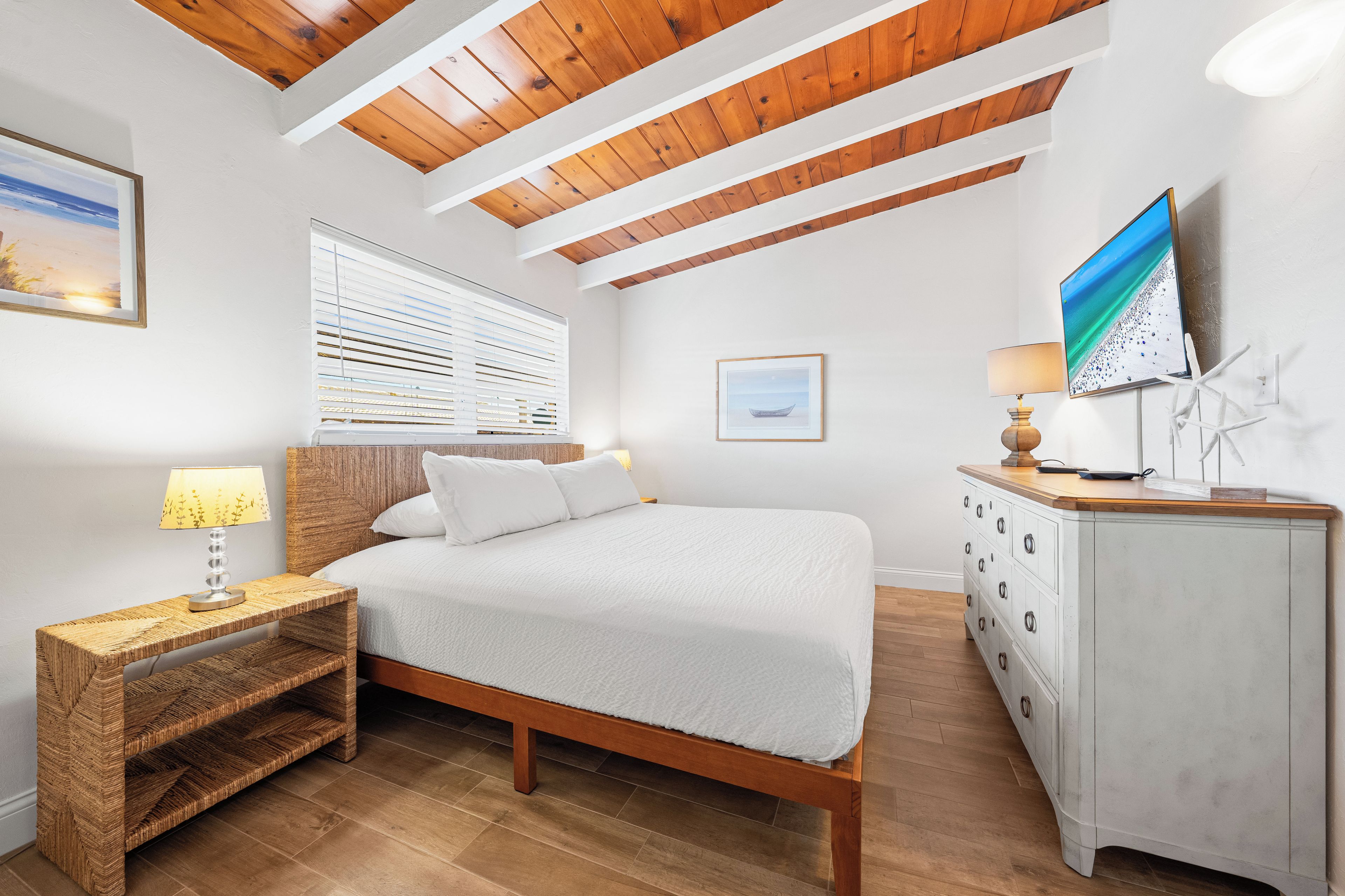 A bedroom with a wooden ceiling, a bed with white bedding, a bedside table with a lamp, a dresser, and a TV on the wall.
