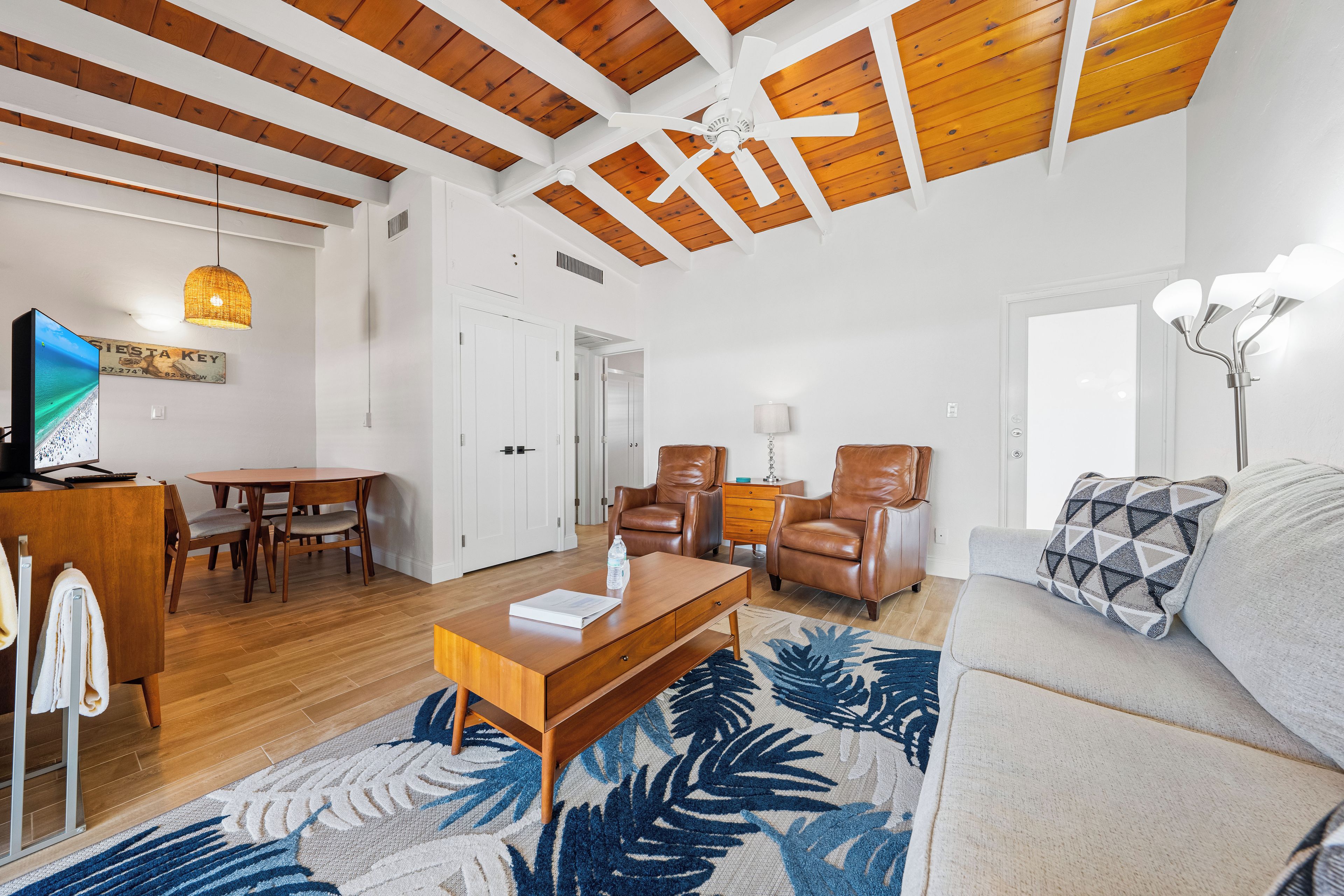 A spacious living room with a high ceiling, wooden beams, a large sofa, a coffee table, a TV, a dining area with a table and chairs, and a patterned rug.