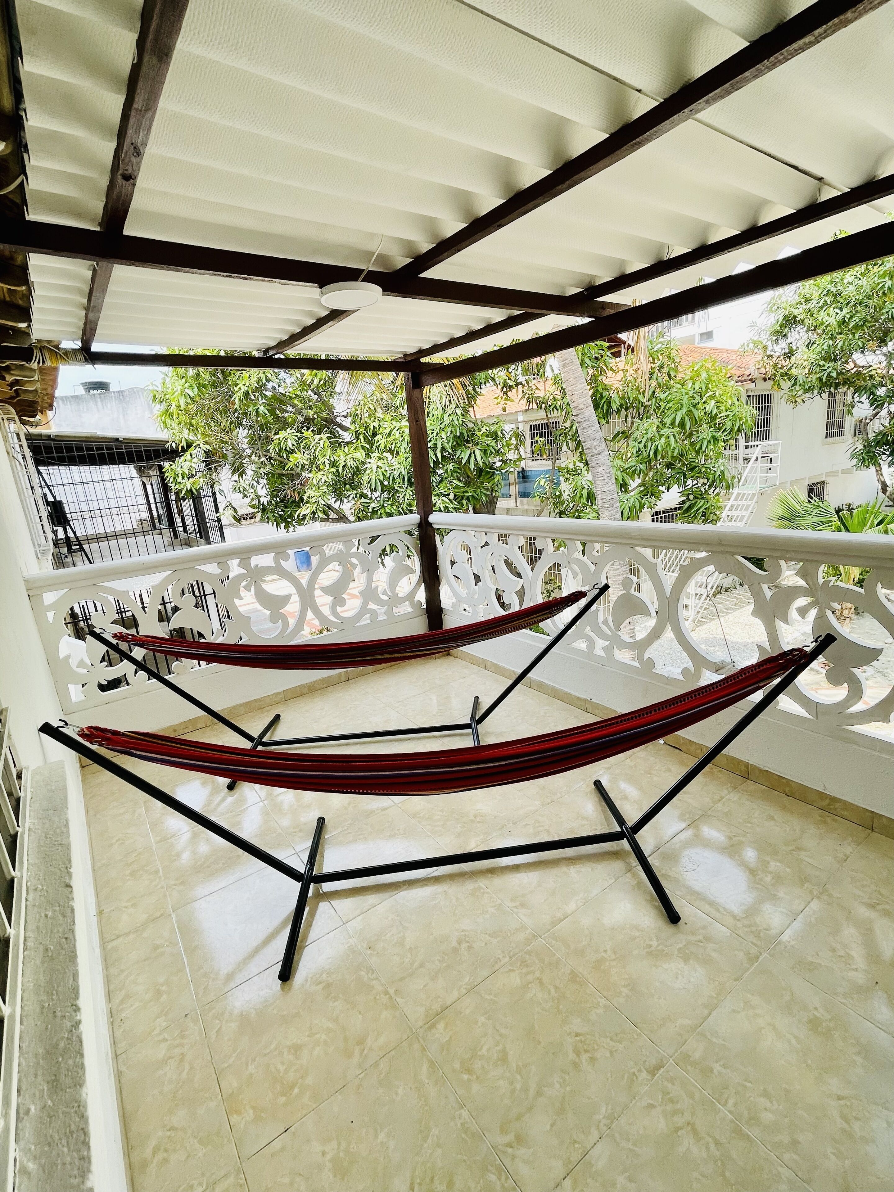 A hammock under a pergola on a tiled balcony with a view of greenery and buildings.