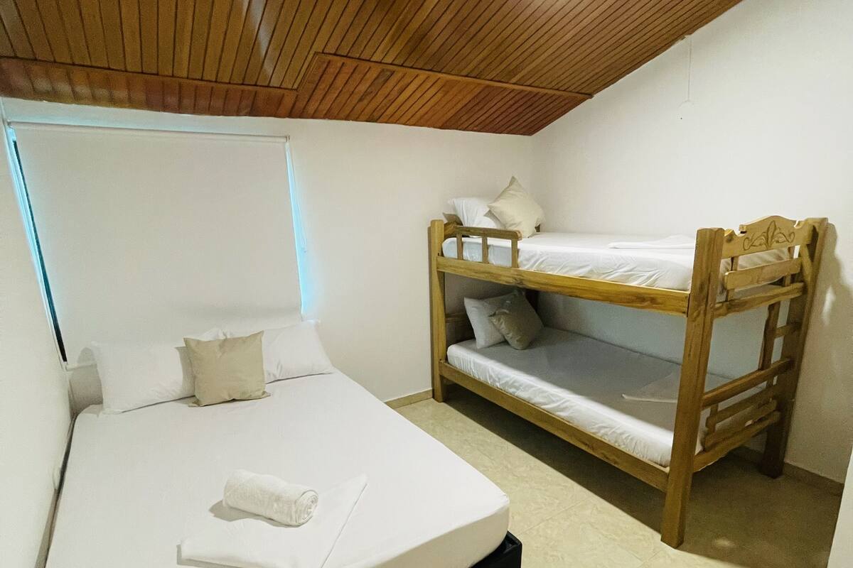 Une chambre avec un lit superposé et un lit simple, tous deux dotés de draps blancs. La chambre a un plafond en bois et des murs blancs.