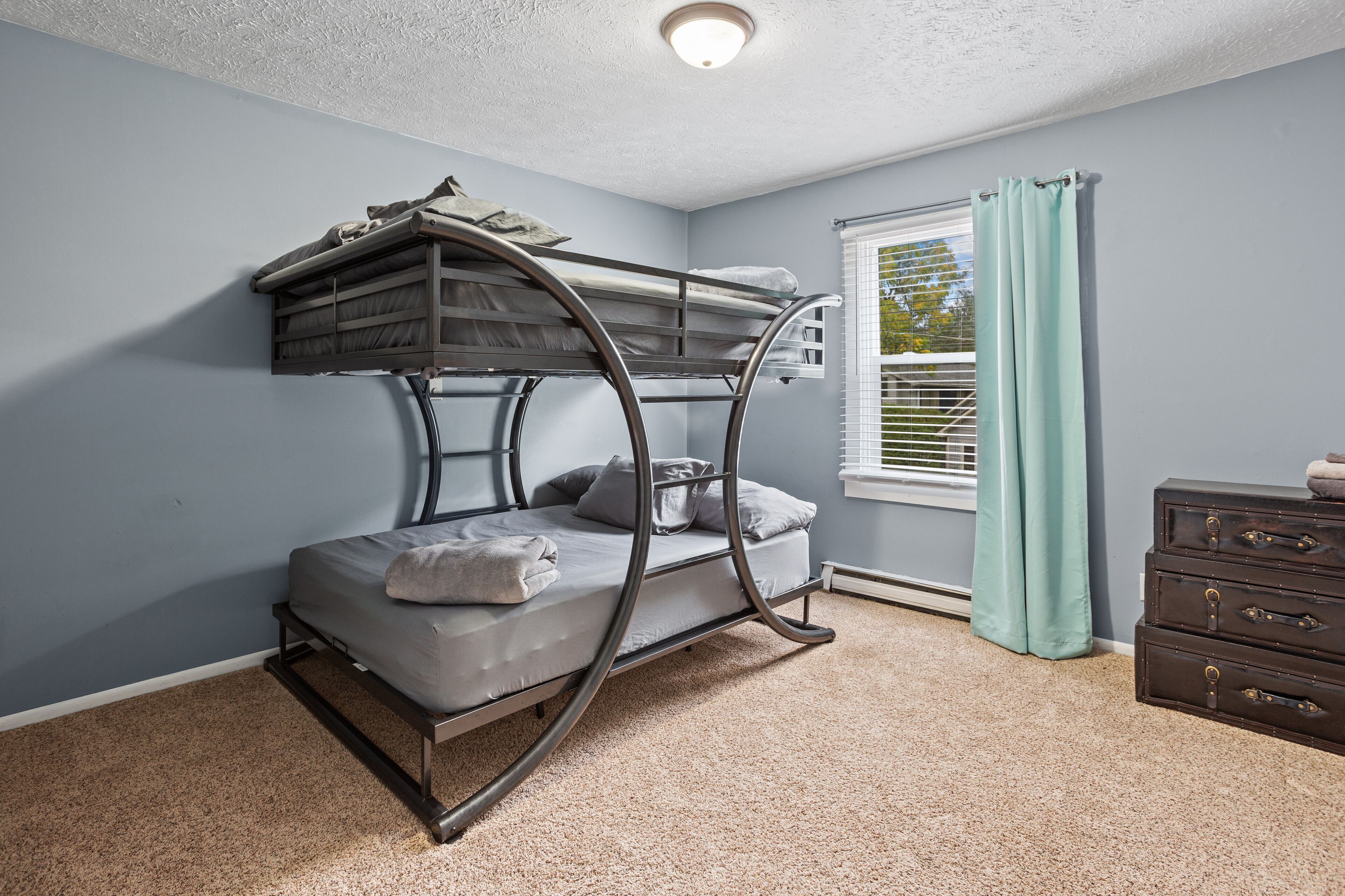 A bunk bed in a room with light blue walls, a window with blinds, and a dresser.