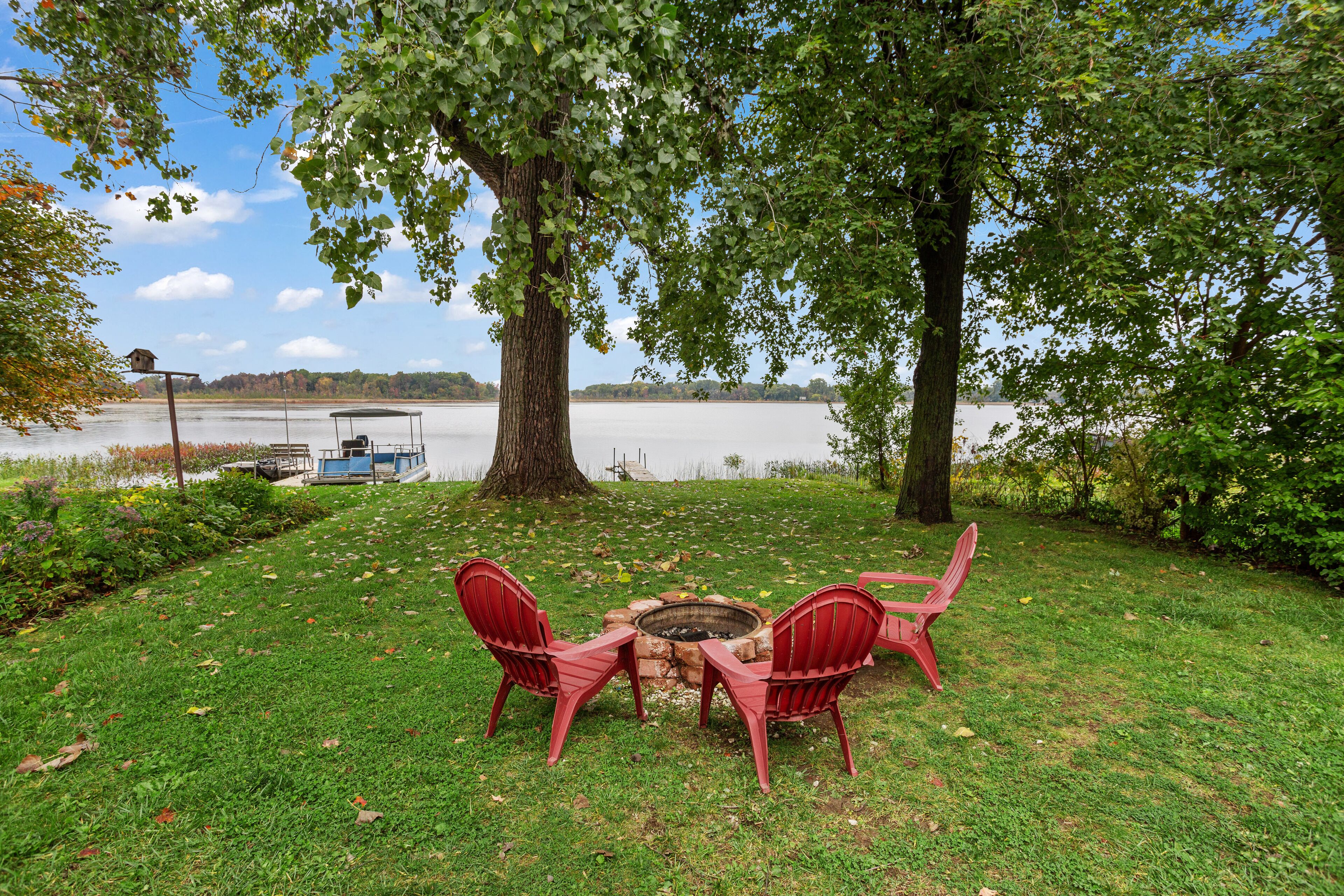 A grassy area with a fire pit and red chairs, overlooking a calm lake with a boat docked in the background.