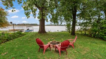 A grassy area with a fire pit and red chairs, overlooking a calm lake with a boat docked in the background.