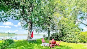 A picnic setup with a table, chairs, and a red umbrella under a large tree by a lake. - Park Lake Getaway (East Lansing)