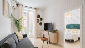 A modern living room with a grey sofa, a wooden TV stand, and a green headboard bed.