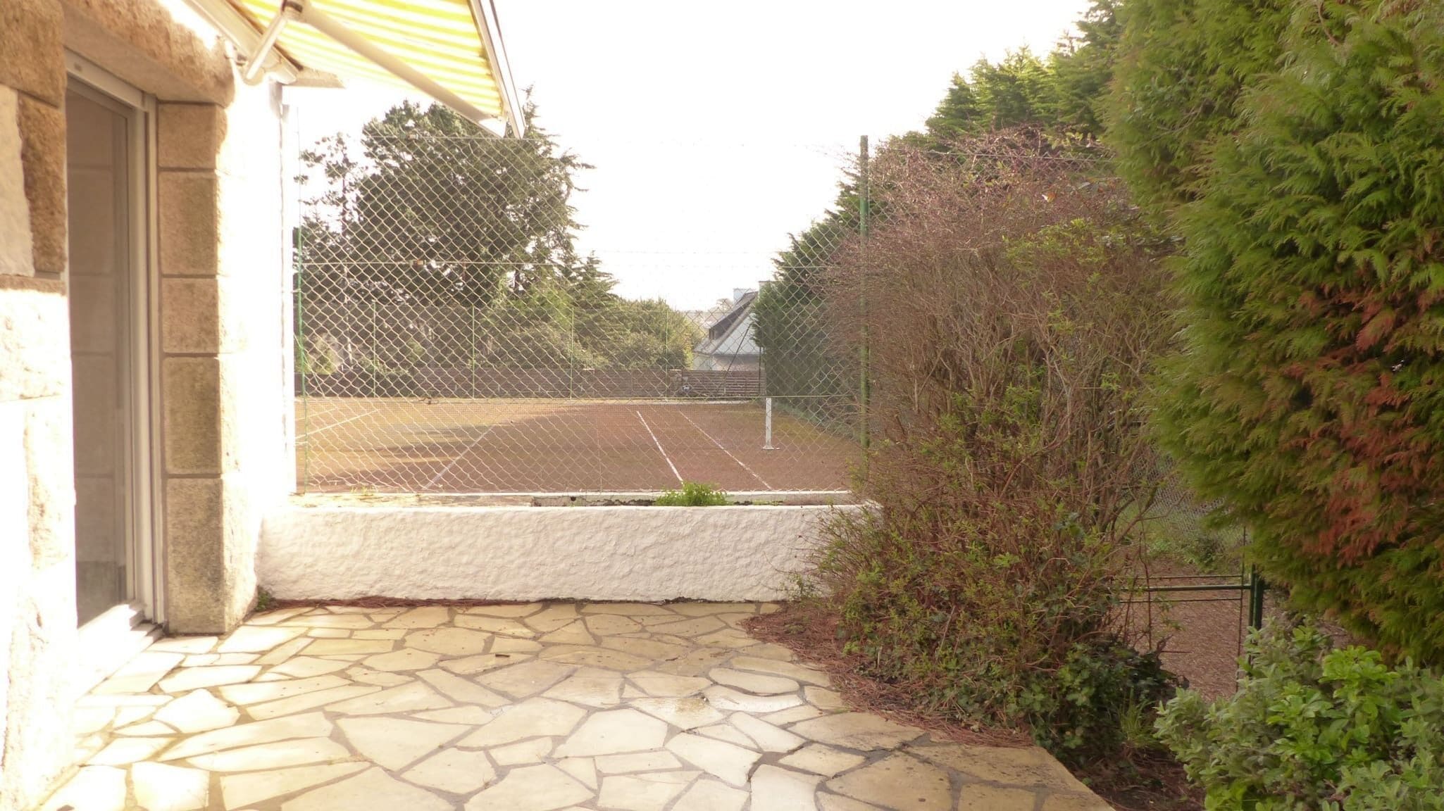 Un patio de piedra que conduce a una cancha de tenis con una cerca de malla metálica y árboles al fondo.
