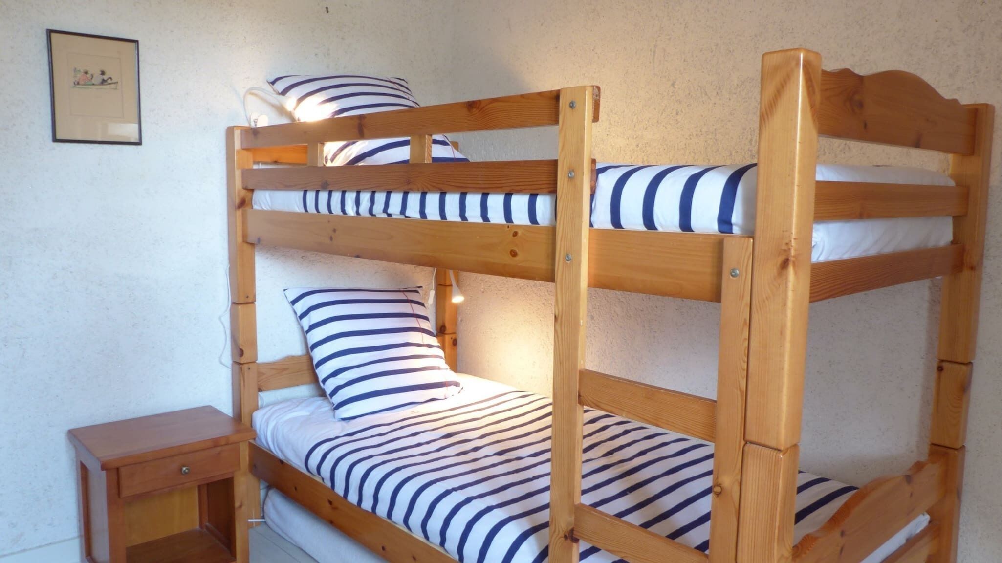 A bunk bed with striped bedding, a bedside table, and a framed picture on the wall.