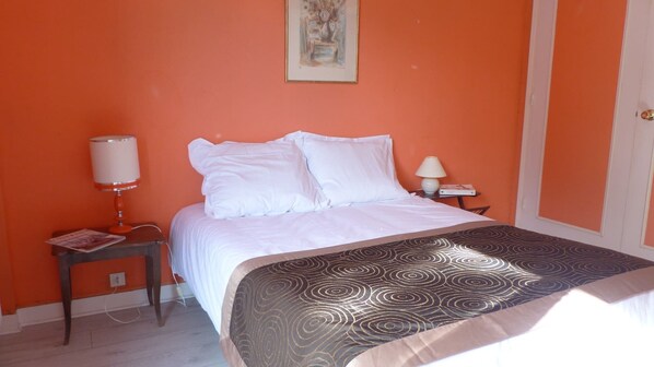 A bedroom with an orange wall, a bed with white bedding, a patterned throw, a nightstand with a lamp, and a painting on the wall.