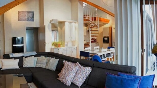 Un espacio habitable moderno estilo loft, con un sofá seccional negro, armarios blancos y una escalera que sube a un nivel superior.