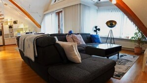Un salón moderno con un sofá gris oscuro, una mesa de centro y una pequeña mesita auxiliar. Hay un ventanal con cortinas de gasa, una planta en maceta y un ventilador sobre un soporte de trípode.