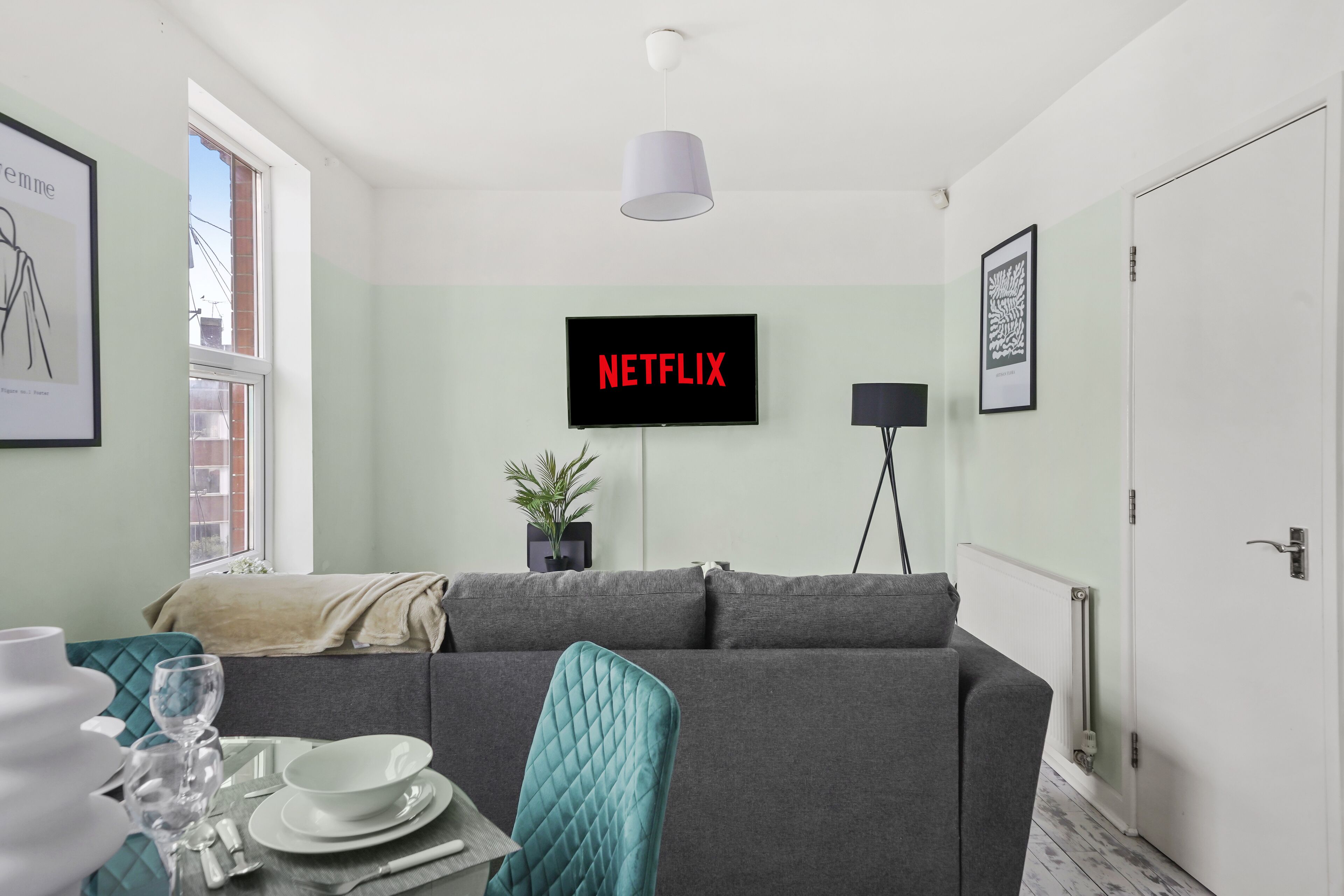 Un salon moderne avec un canapé gris, une table à manger avec des couverts, une plante en pot et un téléviseur affichant le logo « NETFLIX ».
