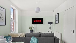 Ett modernt vardagsrum med en grå soffa, ett matbord med bestick, en krukväxt och en tv-skärm som visar "NETFLIX".