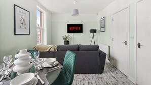 Ett modernt vardagsrum med en grå soffa, ett matbord dukat med tallrikar och glas, en vit väggmonterad tv som visar "NETFLIX", och ett inramat konstverk med titeln "La Femme".