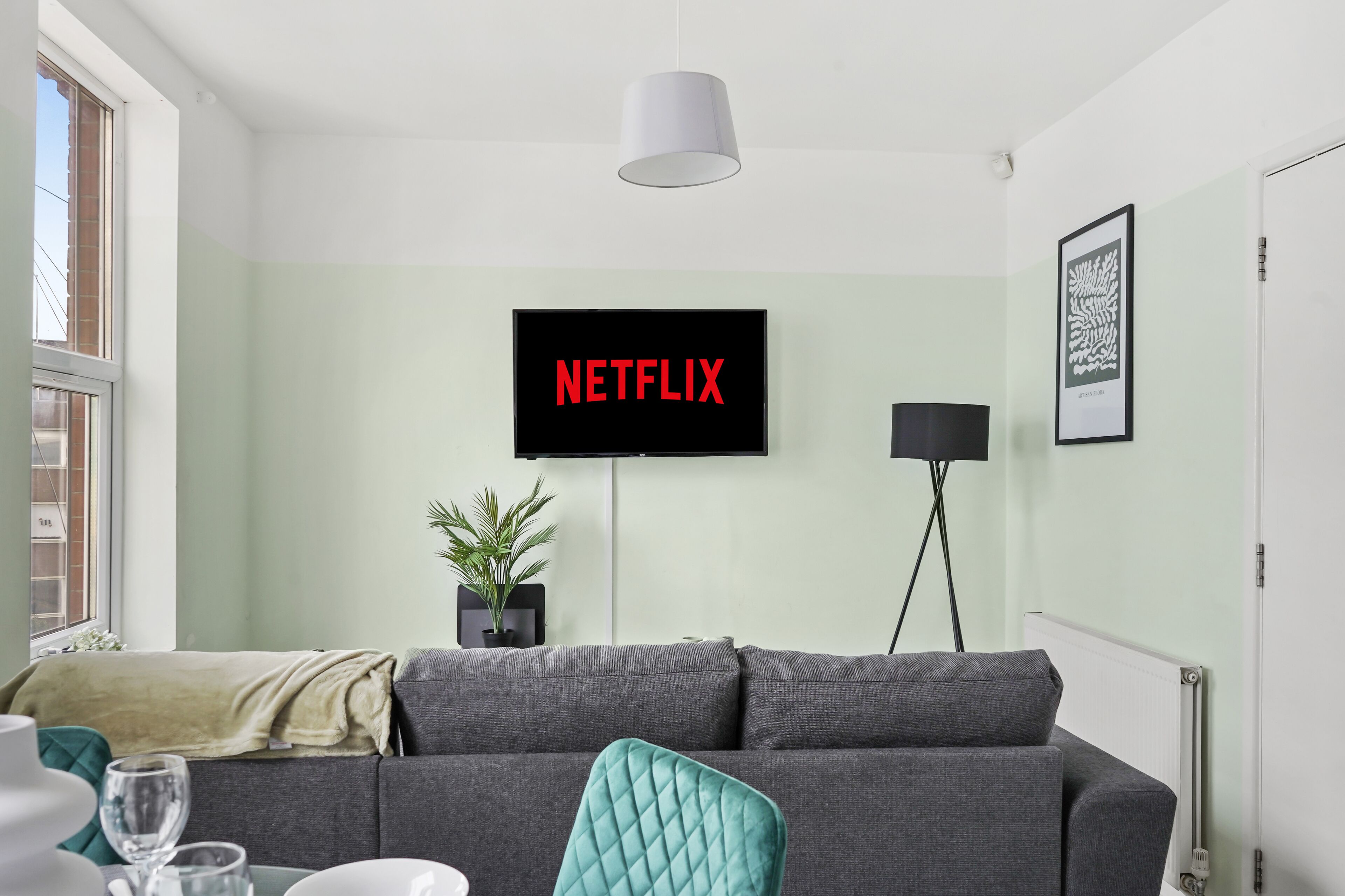 A modern living room with a grey sofa, a teal chair, a potted plant, a black floor lamp, and a TV displaying "NETFLIX".