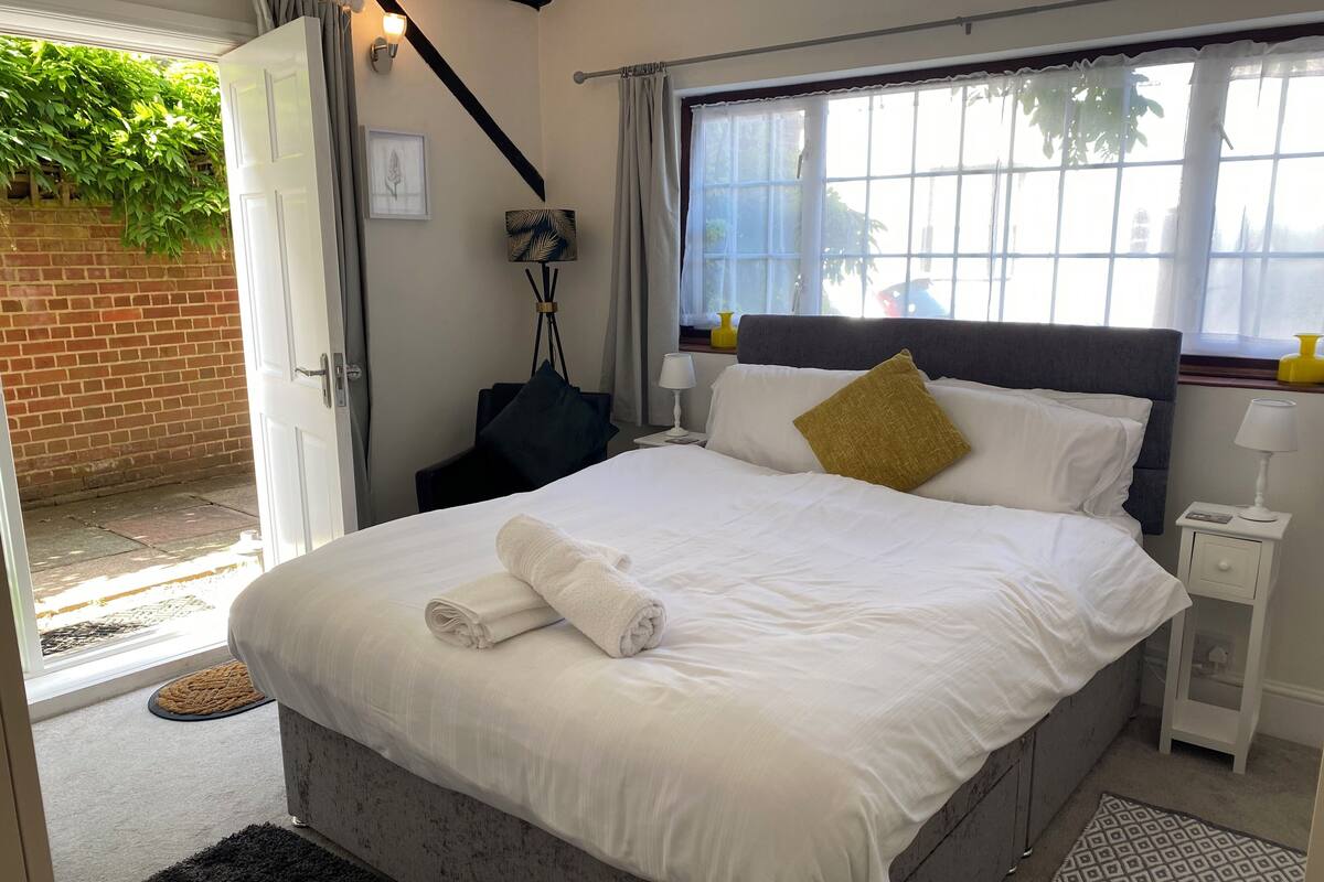 Una cama bien hecha con sábanas blancas y una toalla enrollada, una lámpara de noche y una mesita de noche con una lámpara.