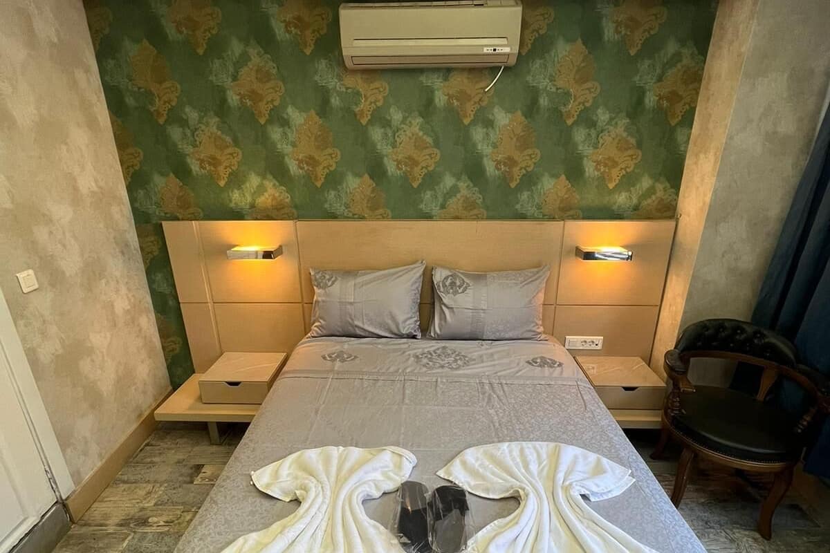 A hotel room with a bed, two bedside tables, an air conditioner, and a chair.