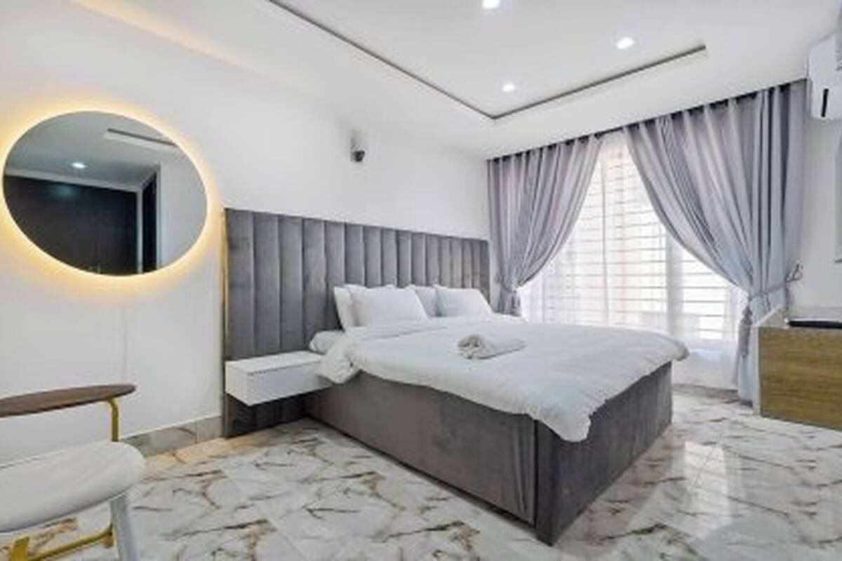 A modern bedroom with a large bed, a bedside table, a round mirror, and a window with curtains.