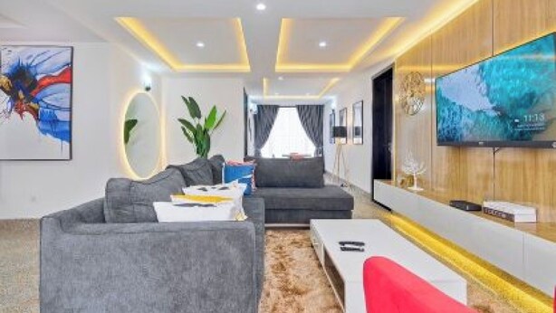 A modern living room with a grey sofa, a red chair, a coffee table, and a TV mounted on the wall. - Tokyo Apartments  (Abuja)