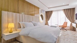 A hotel room with a large bed, a bedside table with a lamp, a desk with a chair, and a large window with curtains. - Tokyo Apartments  (Abuja)