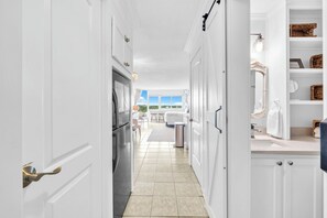 Un pasillo estrecho con armarios blancos, un refrigerador de acero inoxidable y vista a un comedor con mesa y sillas.