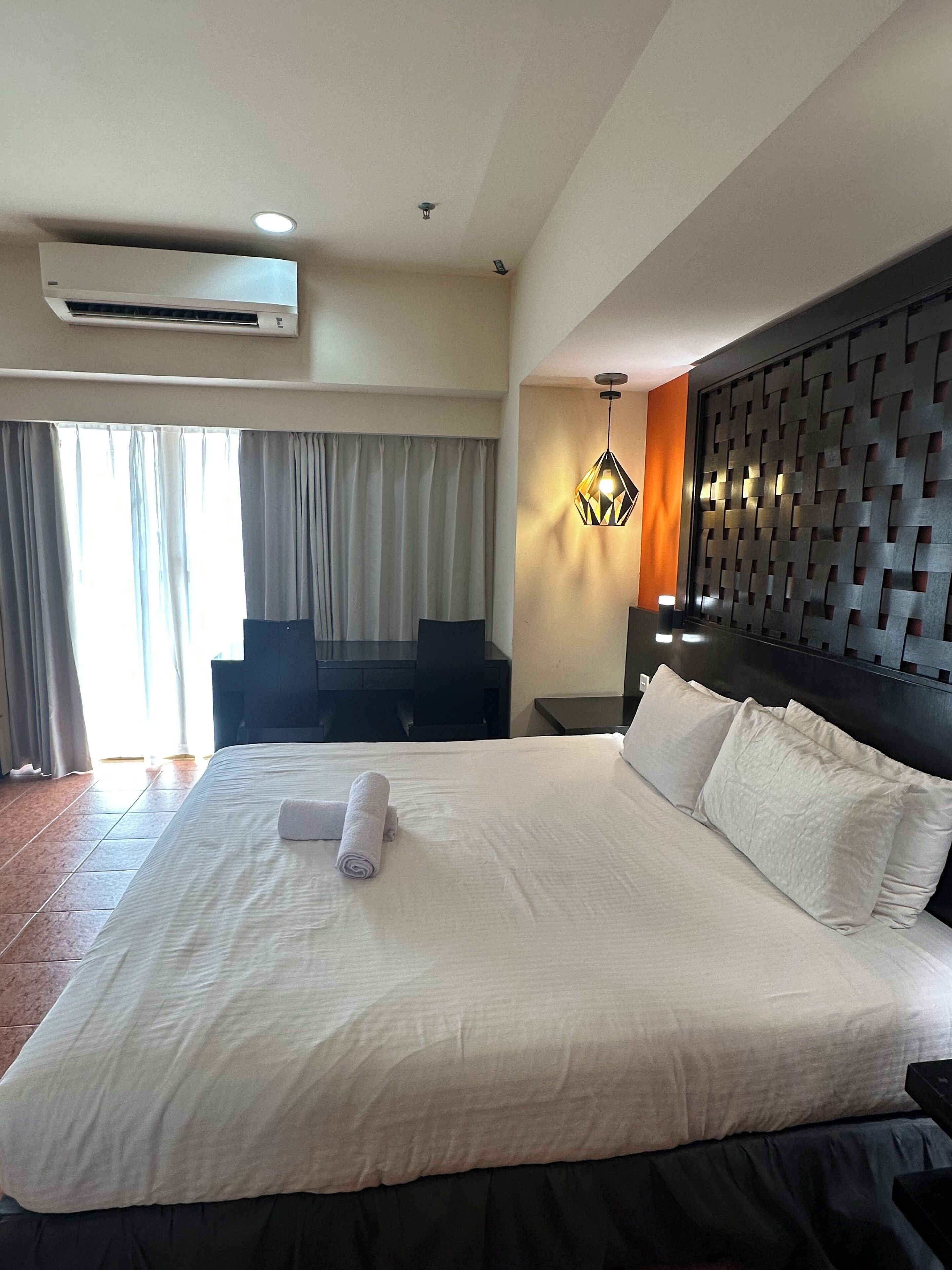 Quarto de hotel com uma cama grande, cabeceira com padrão geométrico, ar condicionado fixado na parede e janela com cortinas.