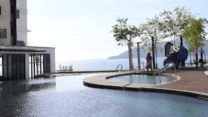 A pool area with a view of the sea, a modern building, and a few trees. - Homesuite' Home at The Shore KK (Kota Kinabalu)