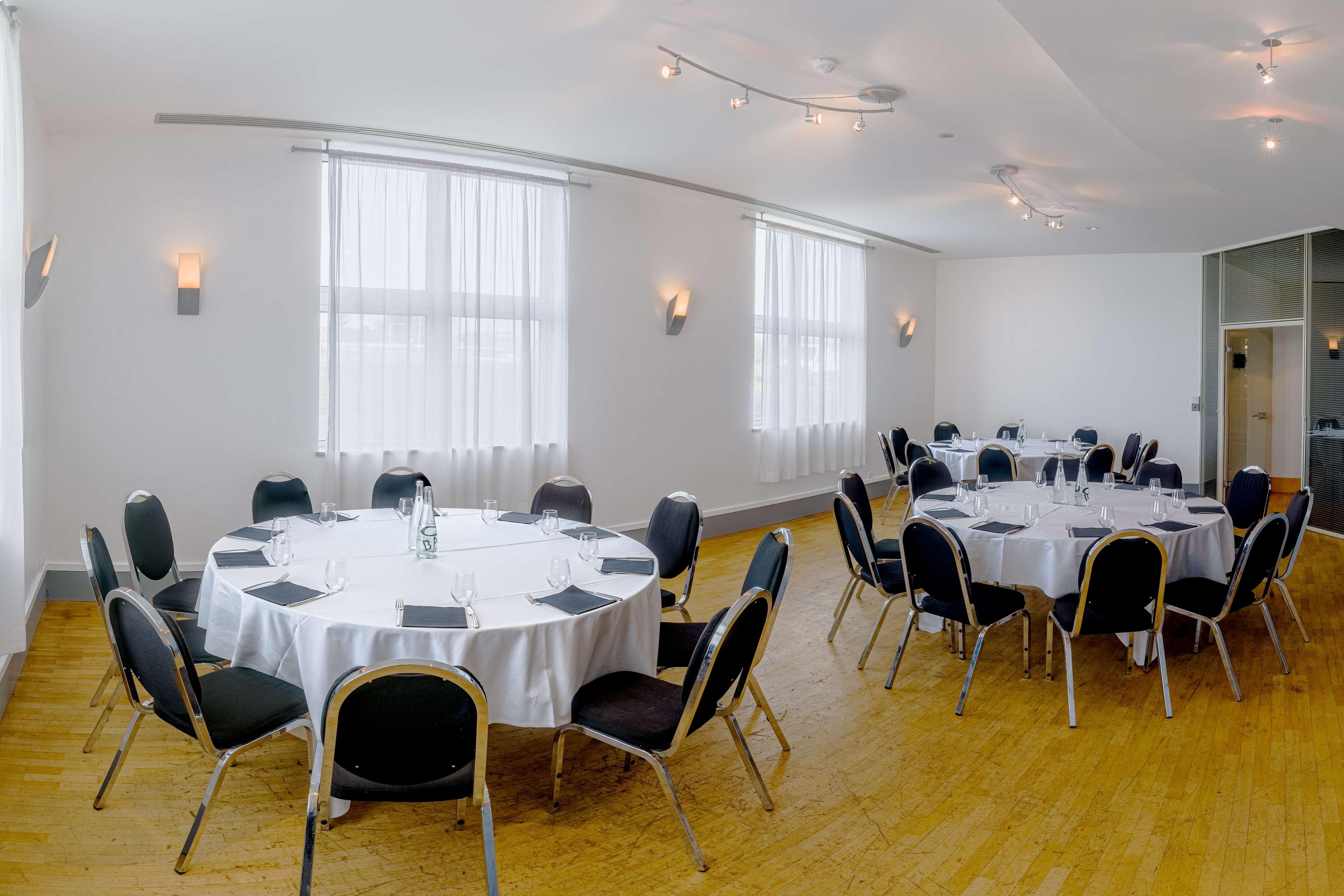 Un salón de conferencias con varias mesas redondas, sillas negras y manteles blancos.