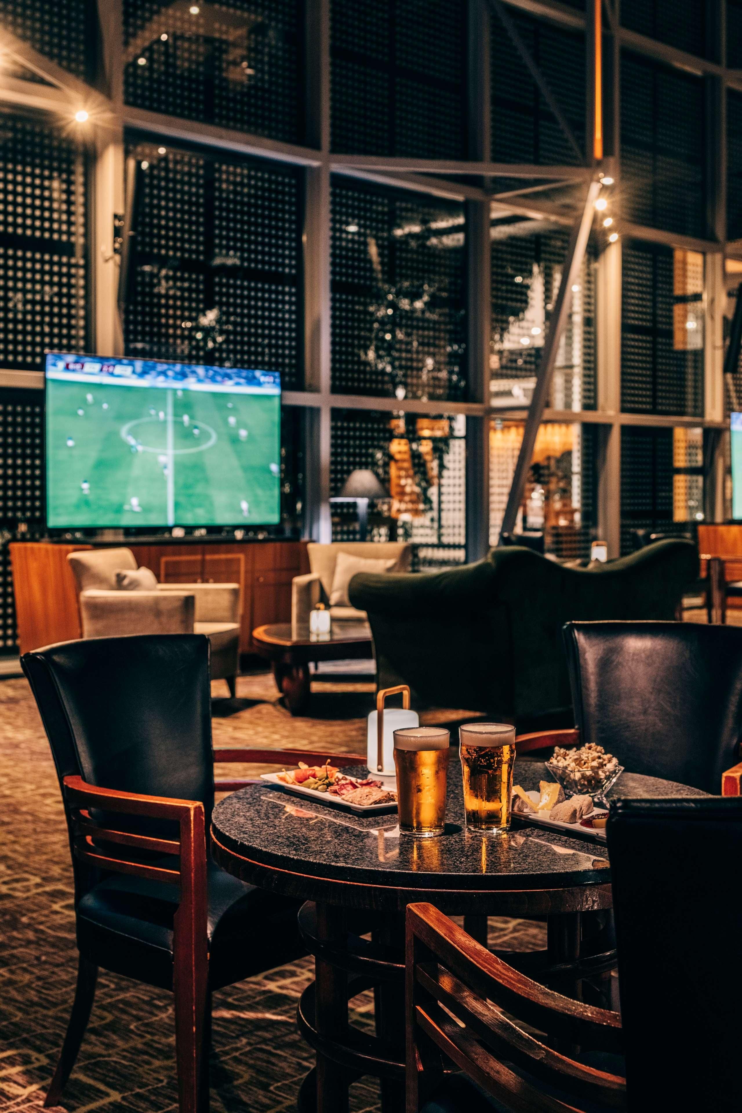 Un área de descanso acogedora con sillones de cuero y una mesa redonda, una pantalla grande mostrando un partido deportivo, y una estantería para vinos al fondo.