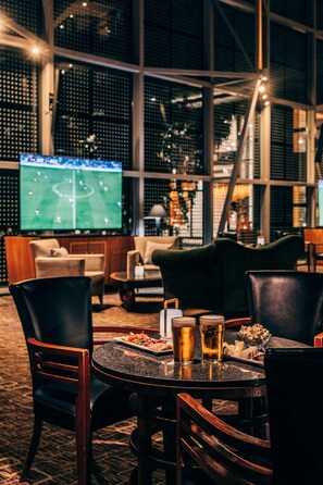 A cozy lounge area with leather chairs and a round table, a large TV screen displaying a sports game, and a wine rack in the background. - Van der Valk Hotel Paris CDG Airport (Roissy-en-France)