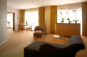 A living room with a sofa, a glass coffee table, a side table, a lamp, a chair, a small table with a chair, a cabinet, and a window with curtains. - Van der Valk Hotel Paris CDG Airport (Roissy-en-France)