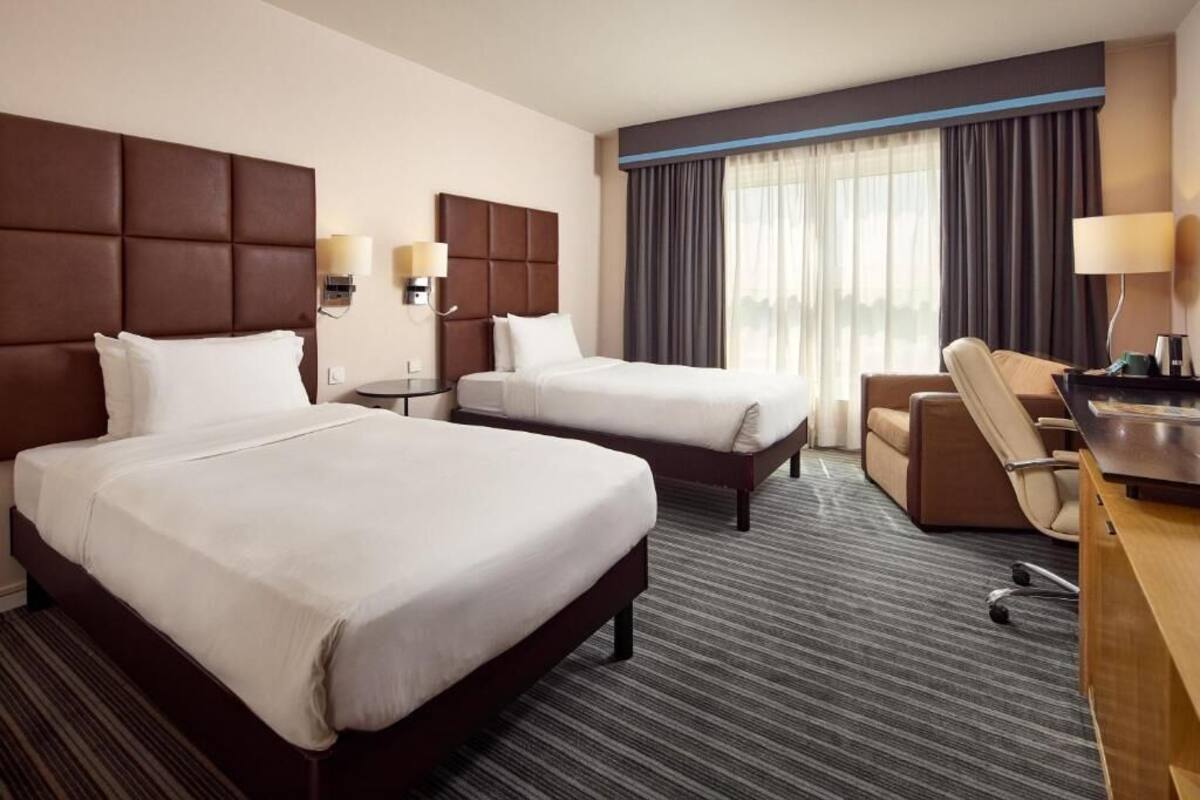A hotel room with two beds, a desk, a chair, and a view of the city through large windows.