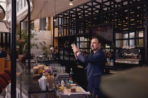 A bar area with a bartender preparing a drink, shelves with bottles, and a coffee machine. - Van der Valk Hotel Paris CDG Airport (Roissy-en-France)