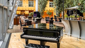 A grand piano in a spacious indoor area with a high ceiling, surrounded by comfortable seating and a large window overlooking a lush garden. - Van der Valk Hotel Paris CDG Airport (Roissy-en-France)
