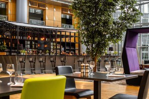 A modern bar area with a high ceiling, wooden paneling, and a large tree. There are bar stools and tables with glasses and bottles. - Van der Valk Hotel Paris CDG Airport (Roissy-en-France)