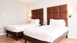 Quarto de hotel com duas camas, cada uma com cabeceira de madeira e roupa de cama branca. Há luminárias de cabeceira e uma pequena mesa com garrafas de água.