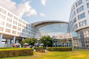 A modern building with a curved glass facade, surrounded by a well-maintained garden with benches and umbrellas. - Van der Valk Hotel Paris CDG Airport (Roissy-en-France)