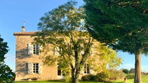 Ein zweistöckiges Steinhaus mit Ziegeldach, umgeben von einem gepflegten Rasen und Bäumen.