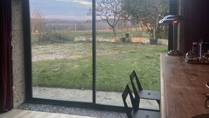 Ein Zimmer mit einem großen Fenster, das Blick auf eine Wiese und Bäume gewährt. Rechts befindet sich ein Holztisch mit Stühlen und einer Lampe.