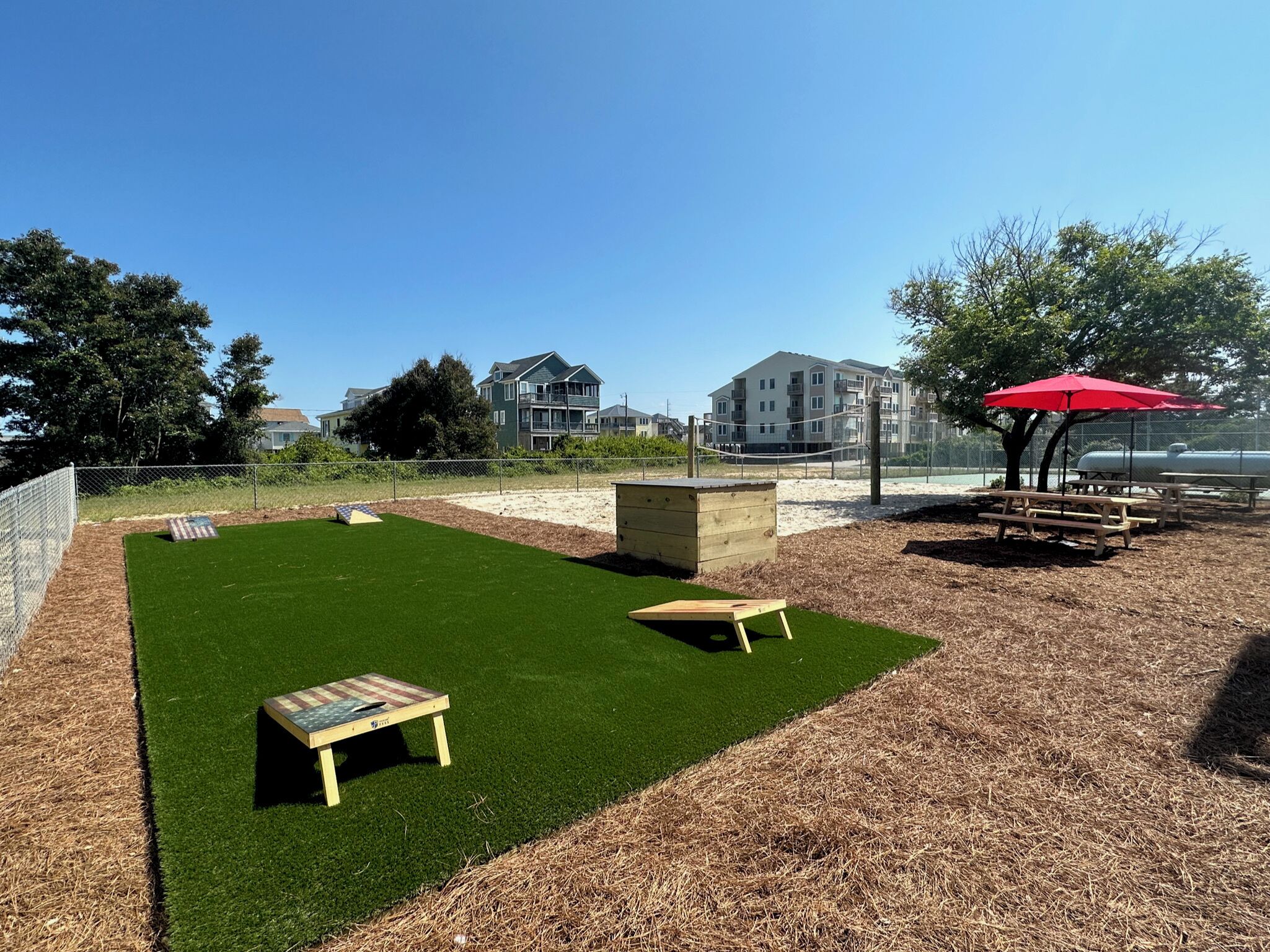 Un jardin avec une pelouse artificielle, un banc en bois, des tables de pique-nique et un parasol rouge.