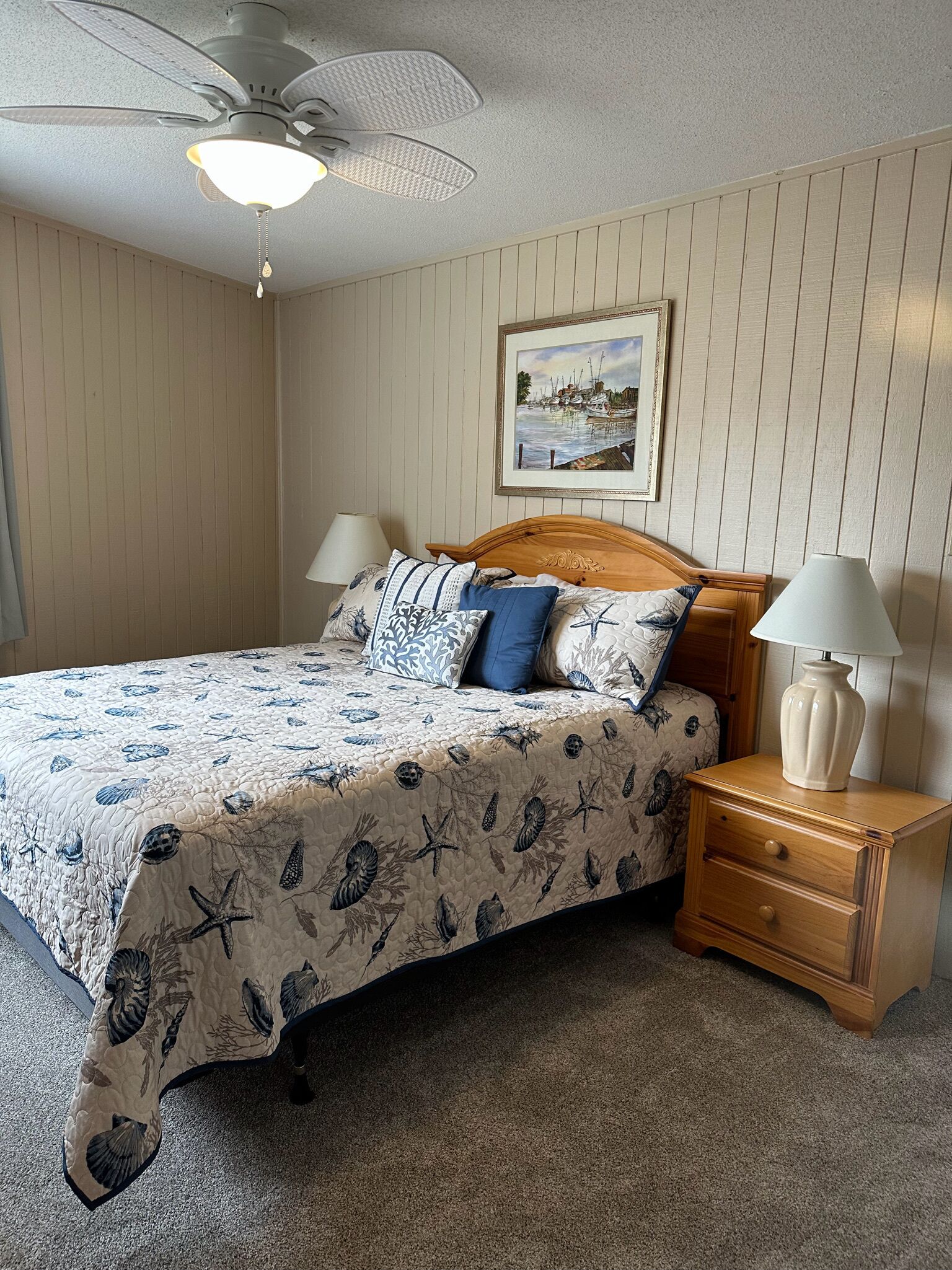 Une chambre à coucher comprenant un lit, des tables de chevet, un ventilateur de plafond, un tableau encadré au mur et un sol recouvert de moquette.