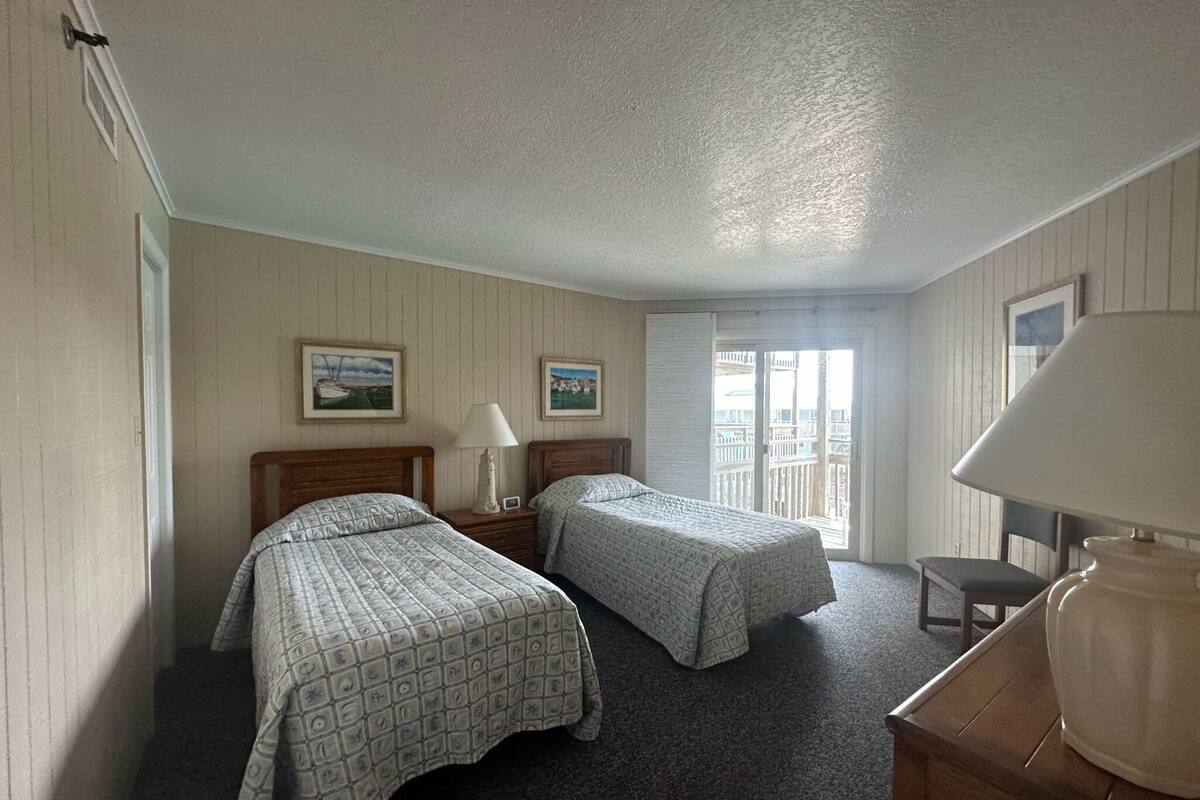 A hotel room with two beds, a desk, a chair, a lamp, and a balcony with a view.