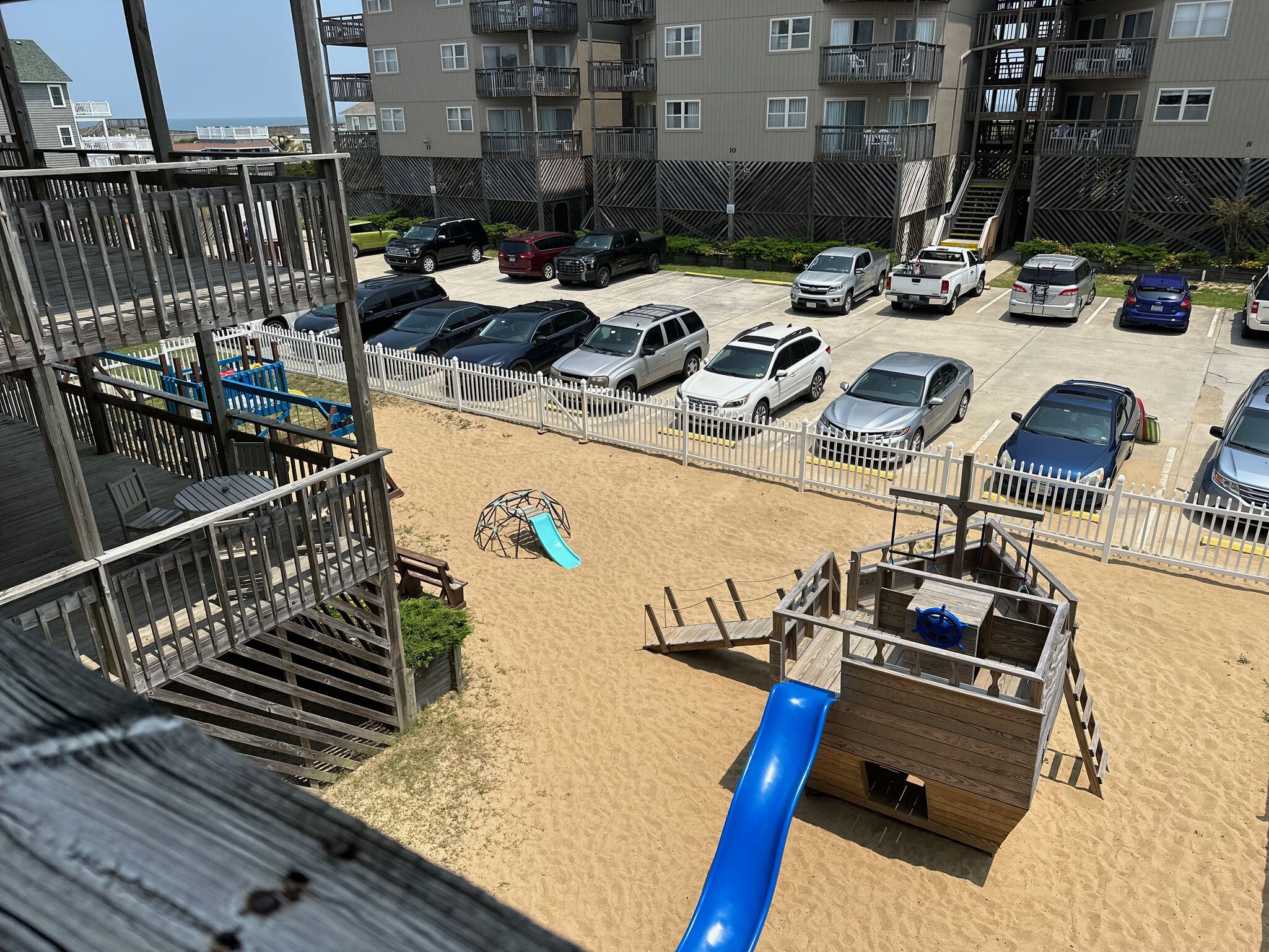 A beachside parking area with multiple cars, a playground with a slide, and a wooden deck.