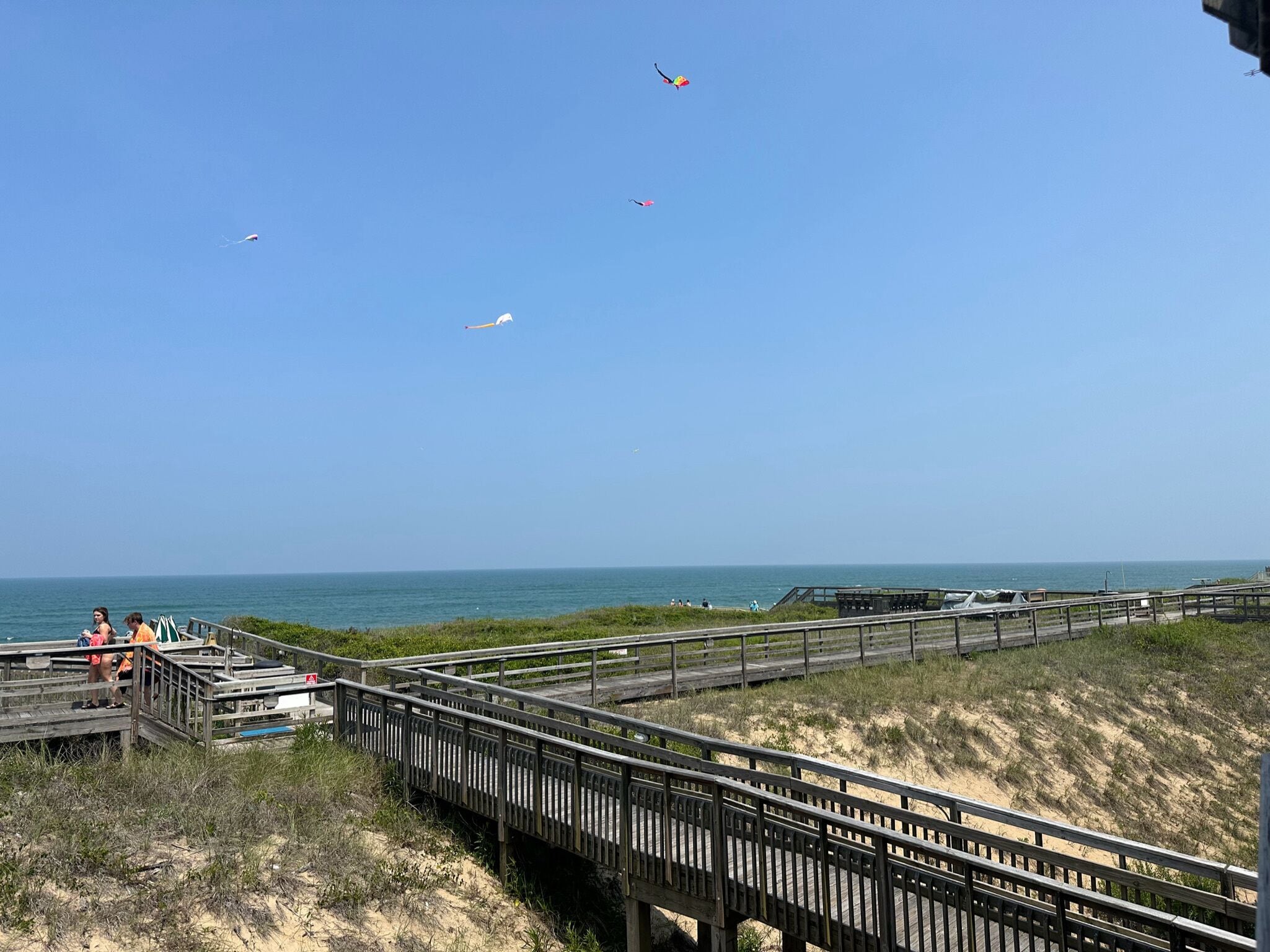 Une promenade en bois qui mène à une plage, avec un ciel dégagé et des cerfs-volants qui volent.